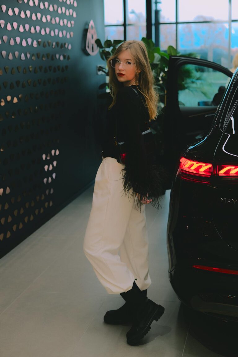 Stylish woman in monochrome outfit stands by a luxury car at a dealership, embracing modern fashion.